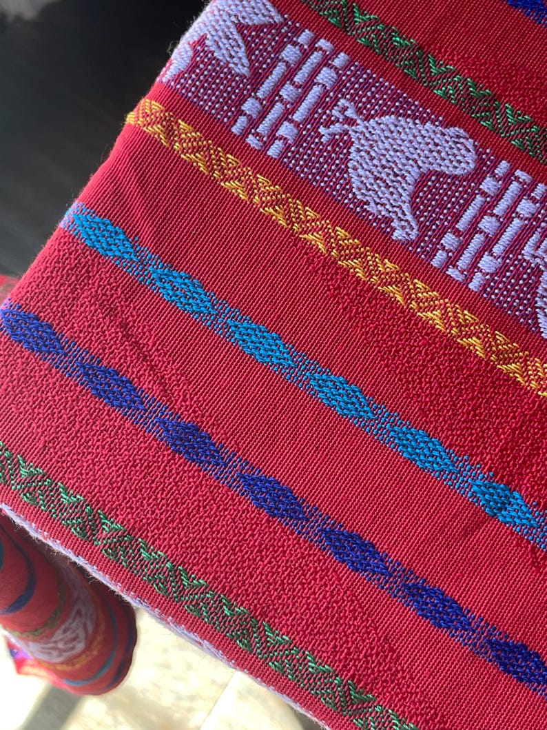 Puede incluir: Textil rojo tejido con rayas coloridas y patrones detallados. El dise&ntilde;o incluye figuras de animales blancos y formas geom&eacute;tricas, con acentos en azul, amarillo y verde. La tela tiene una apariencia texturizada.