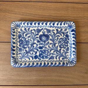 May include: Rectangular ceramic serving tray with a blue and white floral design. The tray features a detailed pattern of stylized flowers and leaves, with a scalloped border. The tray is set against a wooden surface.