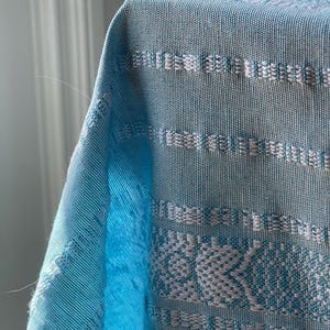 Puede incluir: Primer plano de una tela tejida azul claro con patrones geom&eacute;tricos blancos. La tela tiene una apariencia texturizada y est&aacute; drapeada sobre una superficie. El dise&ntilde;o incluye rayas horizontales y motivos florales, creando un textil decorativo.