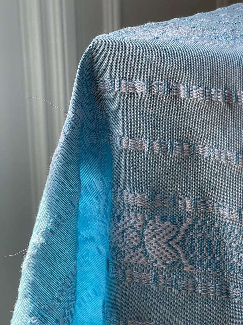 Puede incluir: Primer plano de una tela tejida azul claro con patrones geom&eacute;tricos blancos. La tela tiene una apariencia texturizada y est&aacute; drapeada sobre una superficie. El dise&ntilde;o incluye rayas horizontales y motivos florales, creando un textil decorativo.