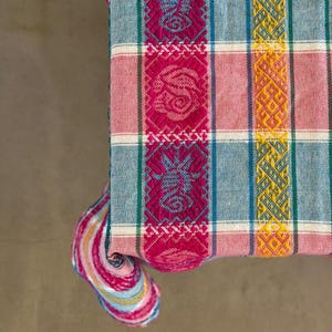 May include: A woven textile featuring a plaid pattern in shades of light blue, pink, and white. The fabric includes vertical bands of vibrant magenta with intricate geometric and floral motifs, alongside yellow bands with detailed diamond patterns.