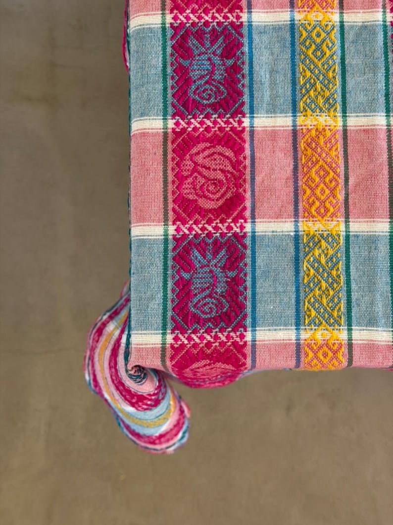 May include: A woven textile featuring a plaid pattern in shades of light blue, pink, and white. The fabric includes vertical bands of vibrant magenta with intricate geometric and floral motifs, alongside yellow bands with detailed diamond patterns.