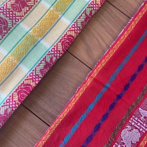 Puede incluir: Dos textiles tejidos coloridos. Uno es amarillo, verde y rosa con un borde floral. El otro es rojo con rayas azules, moradas y blancas y un borde estampado. Los textiles se exhiben sobre una superficie de madera.