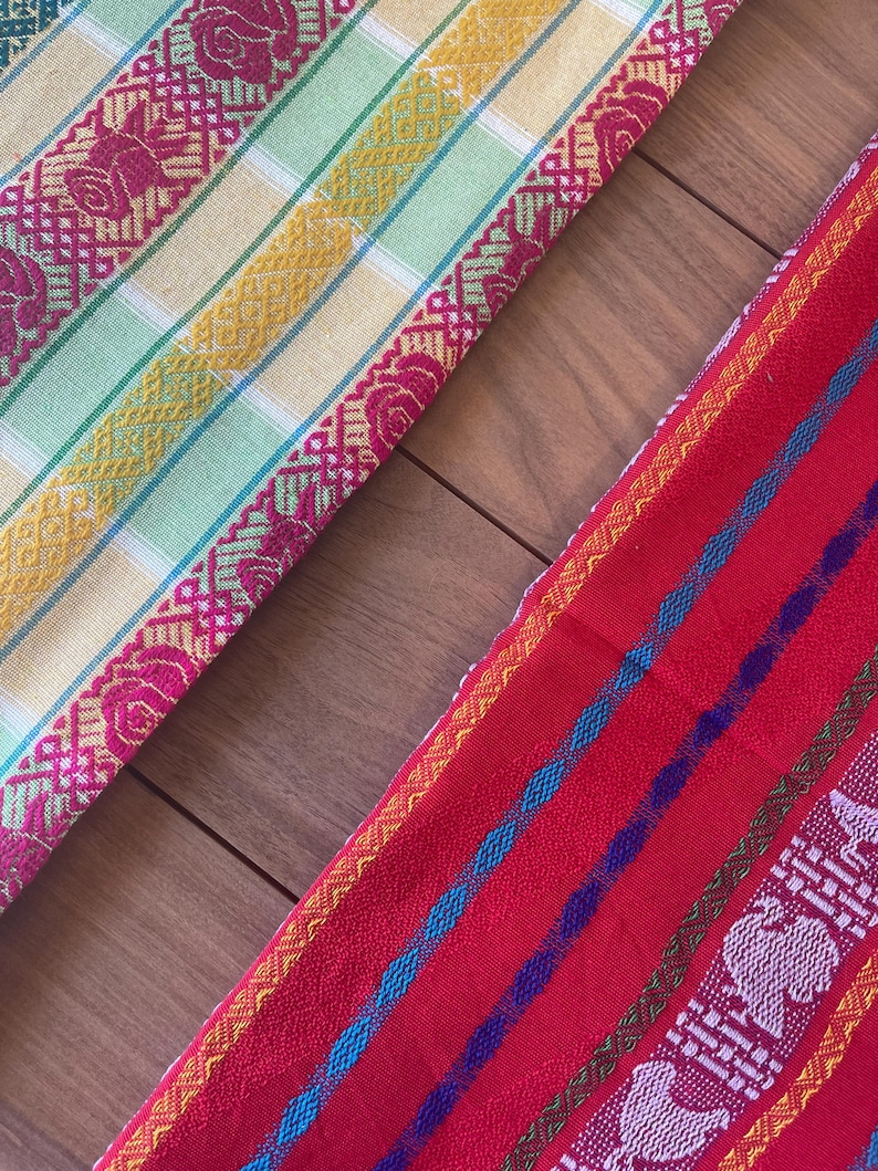 Puede incluir: Dos textiles tejidos coloridos. Uno es amarillo, verde y rosa con un borde floral. El otro es rojo con rayas azules, moradas y blancas y un borde estampado. Los textiles se exhiben sobre una superficie de madera.