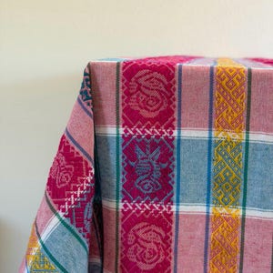 May include: A woven textile featuring a vibrant plaid pattern with vertical stripes in magenta, mustard yellow, and light blue. The fabric displays intricate geometric and floral motifs integrated into the weave, with white accent lines.