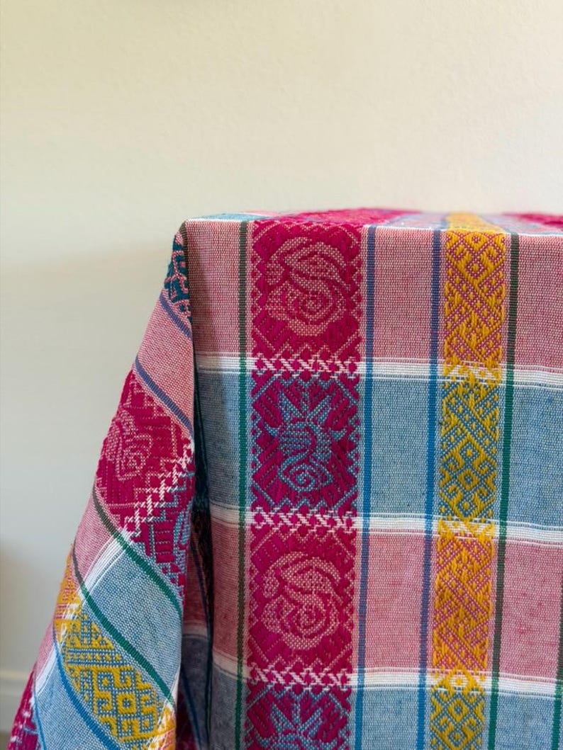May include: A woven textile featuring a vibrant plaid pattern with vertical stripes in magenta, mustard yellow, and light blue. The fabric displays intricate geometric and floral motifs integrated into the weave, with white accent lines.