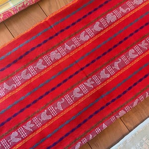 Puede incluir: Un textil rojo vibrante con patrones de aves blancas y geom&eacute;tricos, acentuado por rayas azules, moradas y doradas. La tela parece tejida, con un dise&ntilde;o detallado. Tambi&eacute;n se ven otros textiles en amarillo y azul claro.