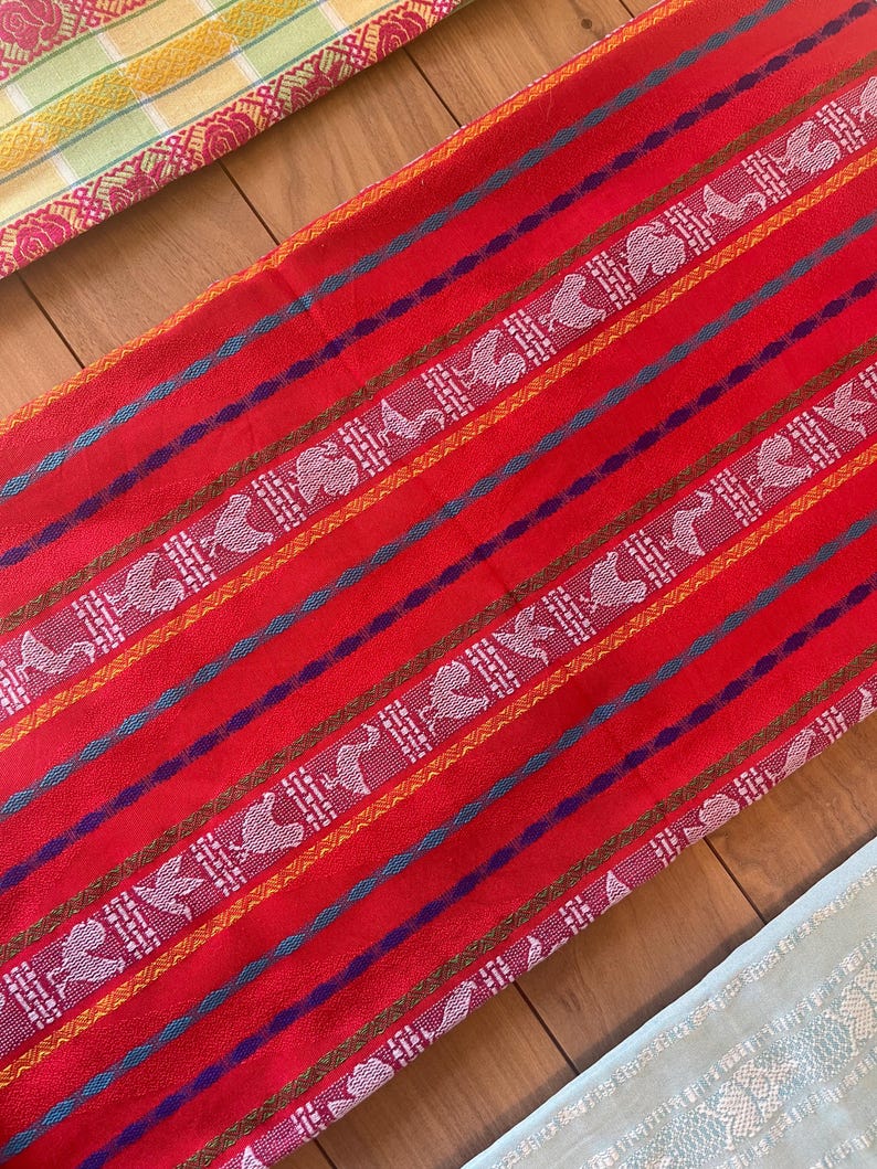 Puede incluir: Un textil rojo vibrante con patrones de aves blancas y geom&eacute;tricos, acentuado por rayas azules, moradas y doradas. La tela parece tejida, con un dise&ntilde;o detallado. Tambi&eacute;n se ven otros textiles en amarillo y azul claro.