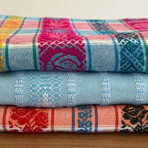 May include: A stack of three folded woven textiles featuring intricate patterns. The top fabric displays a vibrant plaid design in pink, blue, and yellow. The middle fabric is a solid light blue with white stripes. The bottom fabric is orange.