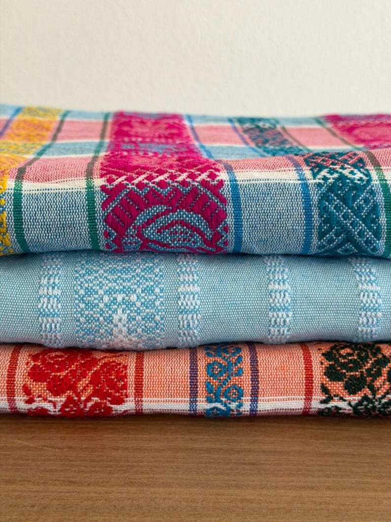 May include: A stack of three folded woven textiles featuring intricate patterns. The top fabric displays a vibrant plaid design in pink, blue, and yellow. The middle fabric is a solid light blue with white stripes. The bottom fabric is orange.