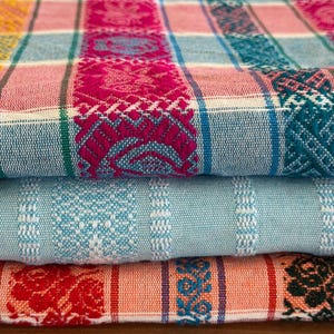 May include: A stack of three folded woven textiles featuring intricate patterns. The top fabric displays a plaid design in magenta, light blue, and white with detailed woven motifs. The middle fabric is light blue with white stripes. The bottom fabric is orange with red and black floral patterns.