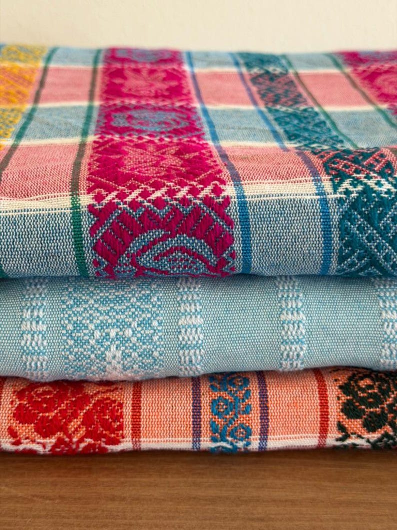 May include: A stack of three folded woven textiles featuring intricate patterns. The top fabric displays a plaid design in magenta, light blue, and white with detailed woven motifs. The middle fabric is light blue with white stripes. The bottom fabric is orange with red and black floral patterns.