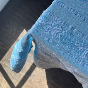 Puede incluir: Un textil tejido azul claro con un patr&oacute;n detallado. La tela est&aacute; drapeada sobre una superficie, con el borde del textil visible. El tejido tiene una apariencia texturizada y el color es un azul p&aacute;lido y suave.