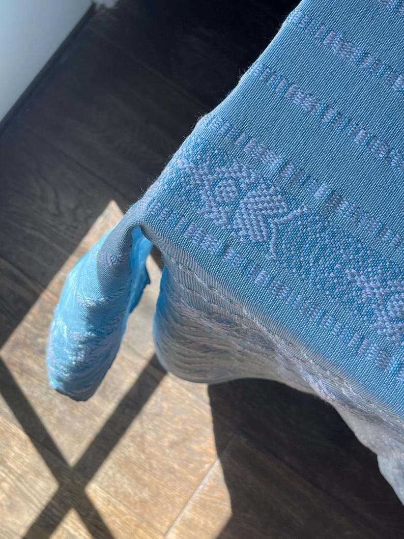 Puede incluir: Un textil tejido azul claro con un patr&oacute;n detallado. La tela est&aacute; drapeada sobre una superficie, con el borde del textil visible. El tejido tiene una apariencia texturizada y el color es un azul p&aacute;lido y suave.