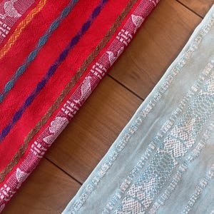 Puede incluir: Primer plano de dos textiles tejidos coloridos. Uno es rojo con rayas y patrones blancos, amarillos, azules y morados. El otro es azul claro con patrones blancos. Los textiles est&aacute;n sobre una superficie de madera.