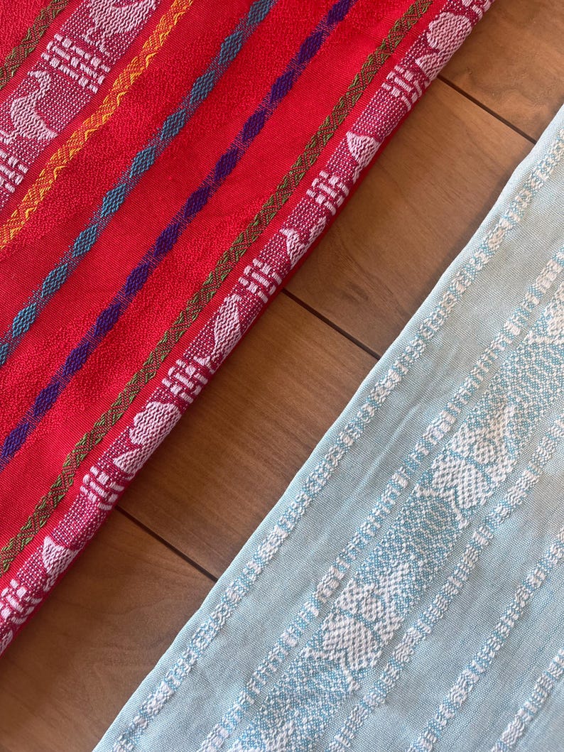Puede incluir: Primer plano de dos textiles tejidos coloridos. Uno es rojo con rayas y patrones blancos, amarillos, azules y morados. El otro es azul claro con patrones blancos. Los textiles est&aacute;n sobre una superficie de madera.