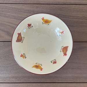 May include: A cream-colored ceramic bowl with a brown rim and chicken illustrations. The bowl features hand-painted chickens in yellow, brown, and red. The bowl is set against a wooden background.