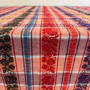 May include: A woven fabric textile featuring a plaid pattern in shades of peach and white, accented by vertical bands of intricate floral motifs in black, bright blue, red, and dark purple, creating a textured and multi-colored surface.