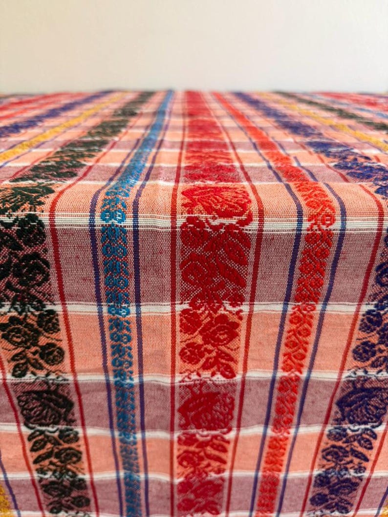 May include: A woven fabric textile featuring a plaid pattern in shades of peach and white, accented by vertical bands of intricate floral motifs in black, bright blue, red, and dark purple, creating a textured and multi-colored surface.