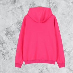 Puede incluir: Una sudadera con capucha de color rosa brillante con capucha a juego y pu&ntilde;os y dobladillo de canal&eacute;. La parte trasera de la sudadera es lisa, con unas peque&ntilde;as salpicaduras de pintura de colores en la manga izquierda. La sudadera con capucha cuelga sobre un fondo gris texturizado.