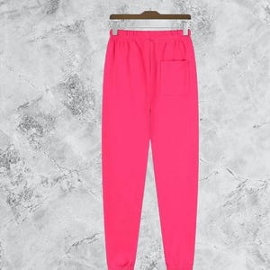 Puede incluir: Pantalones deportivos de color rosa brillante con un bolsillo trasero, colgados de una percha de madera. Los pantalones tienen cintura con cord&oacute;n y tobillos con pu&ntilde;os. El fondo es gris y blanco moteado.