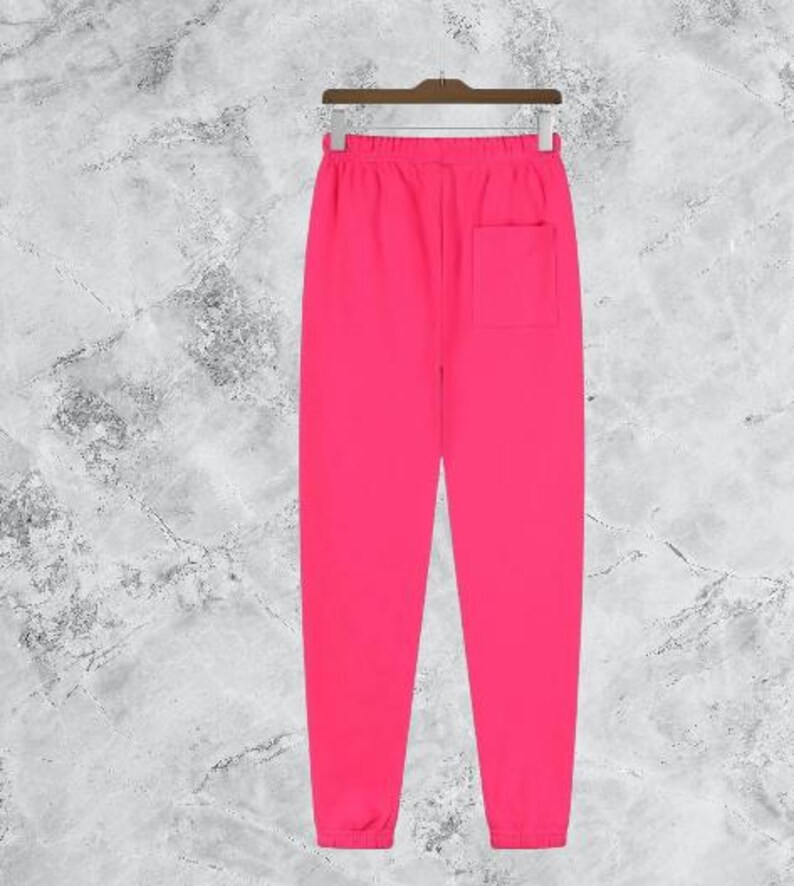 Puede incluir: Pantalones deportivos de color rosa brillante con un bolsillo trasero, colgados de una percha de madera. Los pantalones tienen cintura con cord&oacute;n y tobillos con pu&ntilde;os. El fondo es gris y blanco moteado.