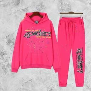 Puede incluir: Conjunto de ch&aacute;ndal rosa brillante que consta de una sudadera con capucha y un pantal&oacute;n de ch&aacute;ndal a juego. La sudadera con capucha presenta un bolsillo delantero y un dise&ntilde;o gr&aacute;fico "Spyder" con una telara&ntilde;a. El pantal&oacute;n de ch&aacute;ndal tiene cintura con cord&oacute;n.