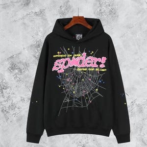 Puede incluir: Sudadera con capucha negra con un dise&ntilde;o gr&aacute;fico que presenta el texto rosa "spider!" y una telara&ntilde;a. El dise&ntilde;o incluye estrellas y el texto "nevermind the punk, here's the slime". La sudadera tiene un bolsillo delantero y una capucha con cord&oacute;n.