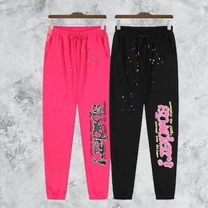 Puede incluir: Dos pantalones de ch&aacute;ndal, uno rosa intenso y otro negro, cuelgan uno al lado del otro. Ambos tienen cintura con cord&oacute;n y la palabra "Spider!" en una fuente decorativa en la pierna izquierda. Salpicaduras de pintura coloridas est&aacute;n dispersas.