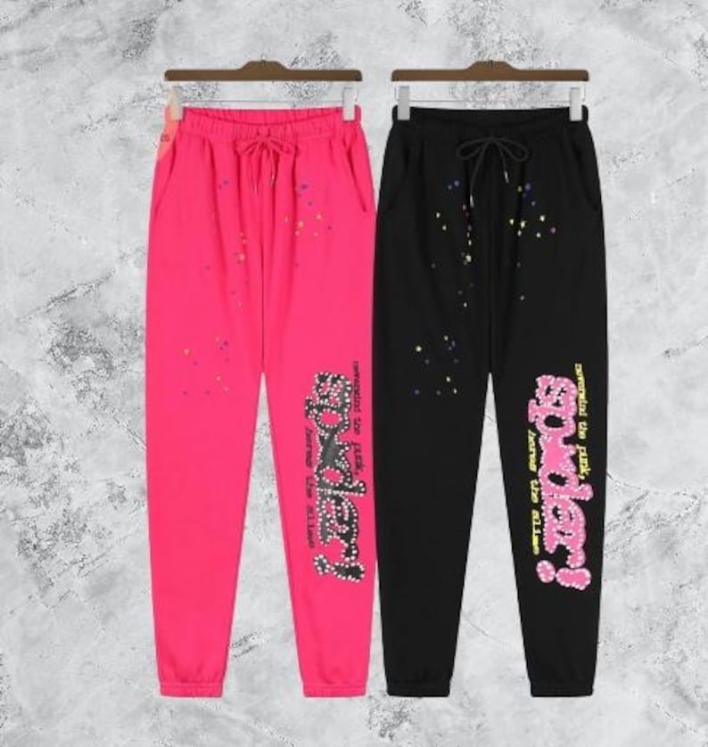 Puede incluir: Dos pantalones de ch&aacute;ndal, uno rosa intenso y otro negro, cuelgan uno al lado del otro. Ambos tienen cintura con cord&oacute;n y la palabra "Spider!" en una fuente decorativa en la pierna izquierda. Salpicaduras de pintura coloridas est&aacute;n dispersas.