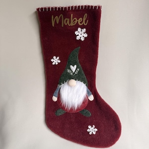May include: A deep red felt Christmas stocking featuring the name Mabel in gold script at the top. The stocking is decorated with three white snowflake appliques and a central gnome character wearing a dark green hat with a white heart design.