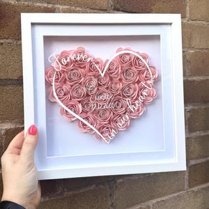 May include: A square white shadow box frame containing a heart-shaped arrangement of light pink paper roses. A white outline of a heart is printed on the glass with the text "Forever in our hearts", the name "Eunice", and the date "12.12.2005".
