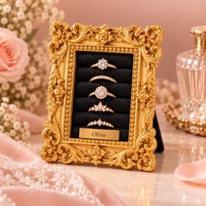 May include: A gold-colored, ornate, rectangular frame with a black velvet insert featuring five horizontal slots for rings. The frame includes a small, rectangular gold plaque at the bottom engraved with the name Olivia.