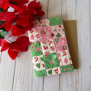 May include: A patchwork Christmas-themed book cover in pink, green, and white, featuring festive patterns like Christmas trees, stockings, and wreaths. Red poinsettia flowers are placed near the book cover on a light wood surface.
