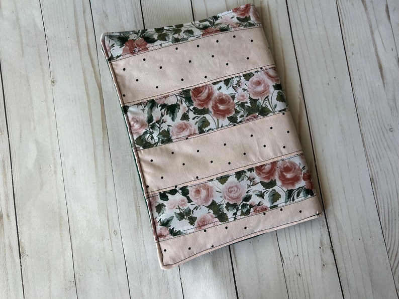 May include: A rectangular book cover with a floral rose pattern and light pink polka dot sections. The cover is made of fabric and has a stitched border. The floral sections feature roses in shades of pink and green.