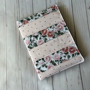 May include: A rectangular, quilted book cover with a floral rose pattern and dotted accents. The cover features alternating stripes of light pink fabric and floral fabric with pink roses and green leaves. Black dots are evenly spaced.