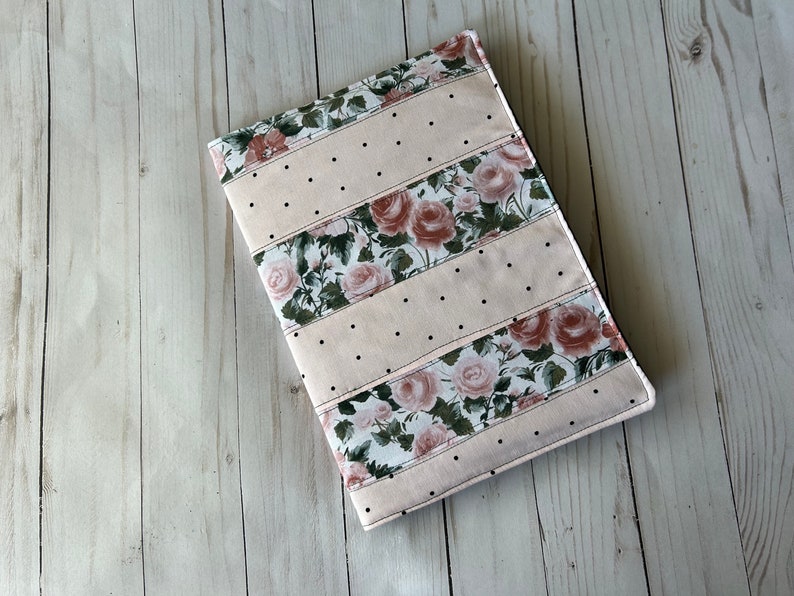 May include: A rectangular, quilted book cover with a floral rose pattern and dotted accents. The cover features alternating stripes of light pink fabric and floral fabric with pink roses and green leaves. Black dots are evenly spaced.