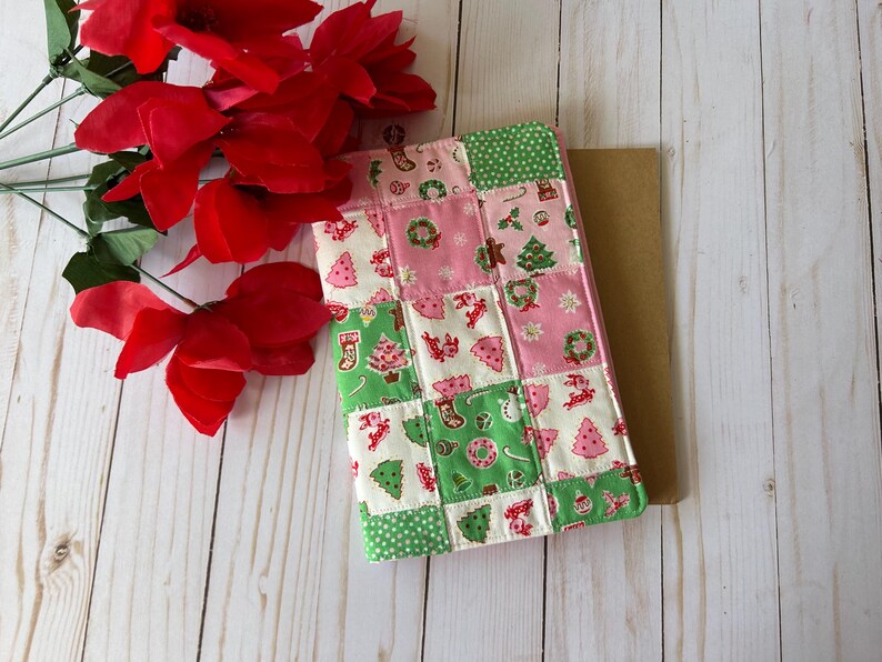 May include: A patchwork Christmas-themed book cover in pink, green, and white, featuring festive patterns like Christmas trees, stockings, and wreaths. Red flowers are placed near the book cover on a light wood surface.