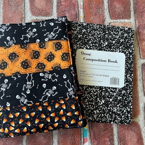 Peut inclure: Une couverture de cahier en tissu avec des bandes horizontales de tissu noir à squelettes blancs, de tissu orange à têtes de chats noirs et de tissu noir à motifs de bonbons, accompagnée d'un cahier à couverture mouchetée noire et blanche.