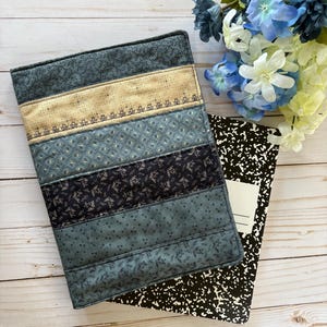May include: A quilted book cover in shades of blue and tan, with a floral pattern, rests atop a black and white composition notebook. The cover is made of fabric strips sewn together. Blue and white flowers are in the background.
