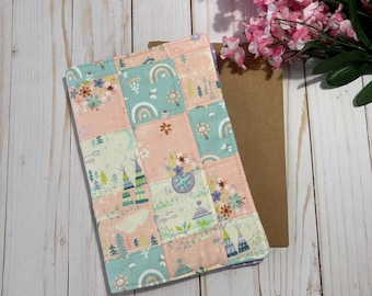 May include: A rectangular fabric book cover featuring a patchwork design of light pink and mint green squares. The pattern includes illustrations of rainbows, teepees, pine trees, flowers, and arrows. The cover is stitched with a quilted texture.