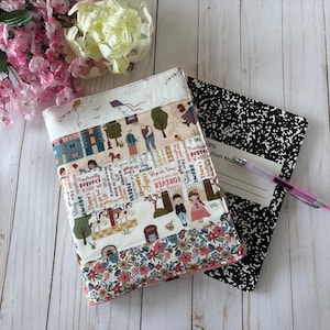 May include: A fabric notebook cover featuring a patchwork design with illustrations of people, trees, and text, layered over a black and white speckled composition notebook with a pen. The cover includes floral patterns and various whimsical character scenes.