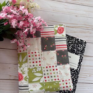 May include: A quilted notebook cover with a patchwork design featuring floral and polka dot patterns in shades of pink, green, and gray. A black and white composition notebook is next to the cover. A bouquet of pink and white flowers is in the background.