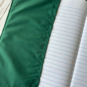 May include: A dark green notebook cover with a stitched edge partially covers a lined notebook. The notebook pages are white with blue horizontal lines. The cover is made of a smooth, solid-colored fabric.
