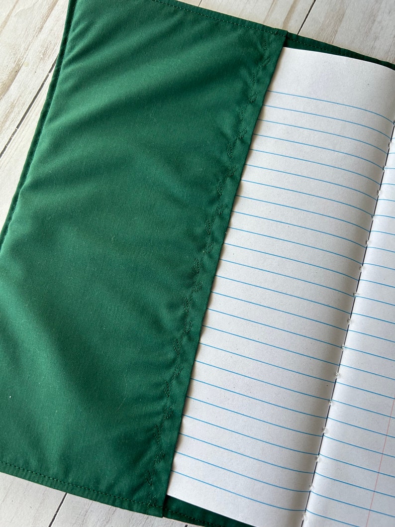 May include: A dark green notebook cover with a stitched edge partially covers a lined notebook. The notebook pages are white with blue horizontal lines. The cover is made of a smooth, solid-colored fabric.