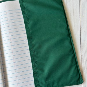 May include: A dark green fabric notebook cover with a stitched leaf design along the spine. The cover is open, revealing lined paper inside. The cover is made of a smooth, solid-colored material.