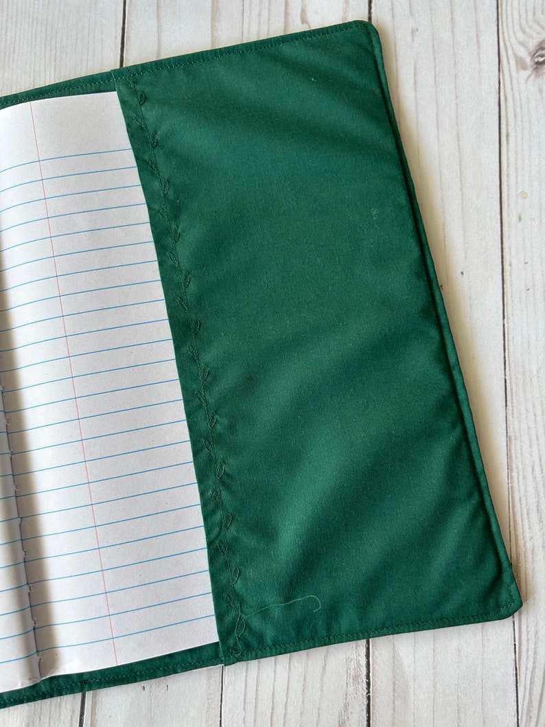 May include: A dark green fabric notebook cover with a stitched leaf design along the spine. The cover is open, revealing lined paper inside. The cover is made of a smooth, solid-colored material.