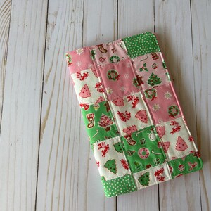 May include: A patchwork Christmas-themed fabric item with pink, green, and white squares. The squares feature festive designs like Christmas trees, wreaths, and stockings. The item is rectangular and appears to be a small case or cover.