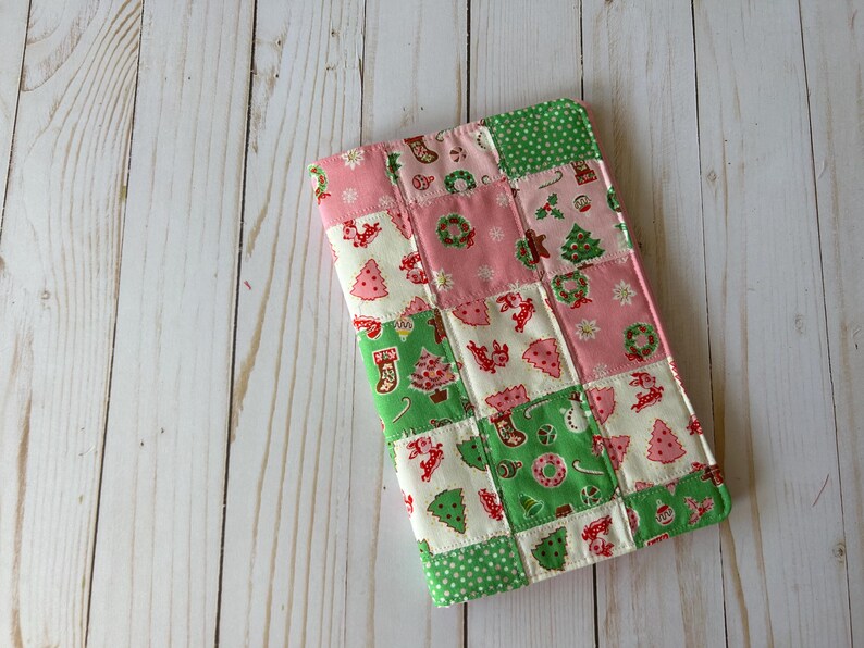 May include: A patchwork Christmas-themed fabric item with pink, green, and white squares. The squares feature festive designs like Christmas trees, wreaths, and stockings. The item is rectangular and appears to be a small case or cover.