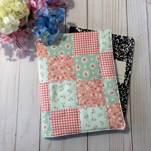 May include: A quilted notebook cover with a patchwork design in shades of red, pink, and mint green. The cover features floral and gingham patterns. A composition notebook is partially visible behind the cover, with a bouquet of flowers in the background.